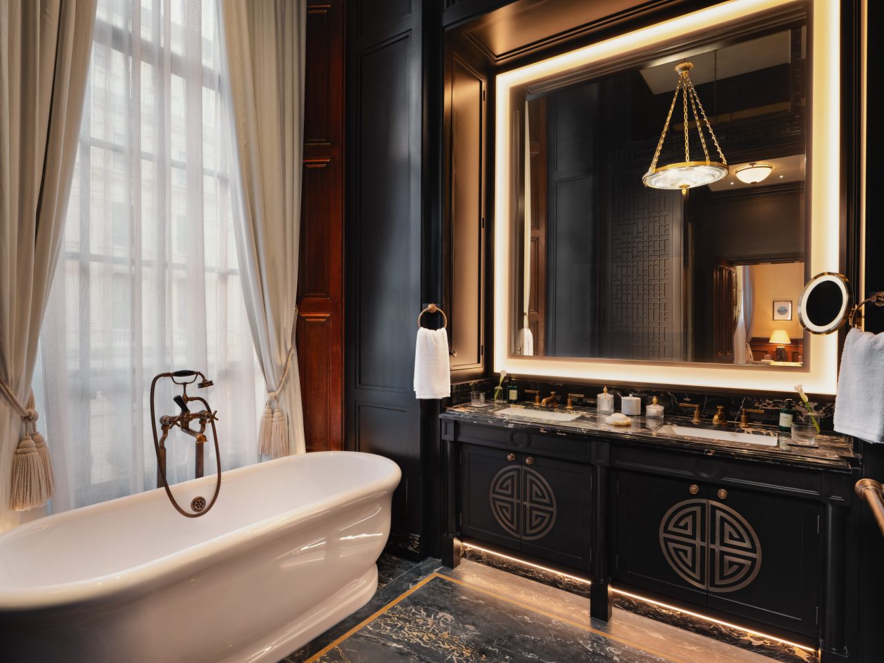 salle de bains à l’ambiance tamisée, avec une grande baignoire blanche devant des rideaux drapés et un miroir cerclé d’une lumière éclatante.